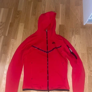 Röd Nike hoodie med dragkedja - Röd hoodie från Nike med hel dragkedja framtill och svart Nike-logga på bröstet. Jackan har huva, långa ärmar och en snygg detalj med svart dragkedja på ena ärmen. 