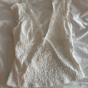 Vit spetsblus med djup v-ringning - Superfin vit blus i spets med djup v-ringning och volangkantade axelband. Blusen har öppen rygg med knäppning och är täckt av blommigt spetsmönster. 