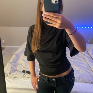 Svart topp från Gina Tricot - Säljer en svart topp från Gina Tricot med lös passform upptill och tightare midja. Toppen har halvlånga, vida ärmar och hög hals. Materialet är slätt och har en lätt glansig finish som ger en stilren look.