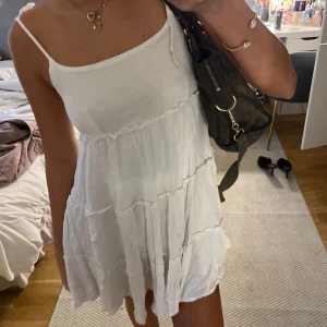 Vit somrig kortklänning med volanger - Superfin vit kortklänning med tunna axelband och flera volanglager. Klänningen har en luftig och flowy passform, perfekt för varma dagar. Materialet känns lätt och svalt mot huden. Enkel och stilren design som ger en fräsch vibe.