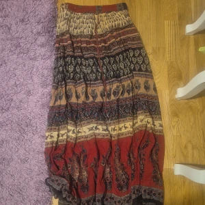 Boho långkjol från Lipo Lipo - Långkjol i boho-stil från Lipo Lipo med vackert mönster i rött, beige och svart. Kjolen har snörning i midjan med små metallpärlor och är tillverkad i ett lätt, flowy material. Perfekt för dig som gillar unika detaljer och sköna vibes.