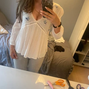 Vit blus med broderade blommor Flash Woman - Superfin vit blus från Flash Woman med v-ringning, broderade blå blommor och spetsdetaljer. Lätt och luftig med trekvatsarmar. Perfekt för vår och sommar.