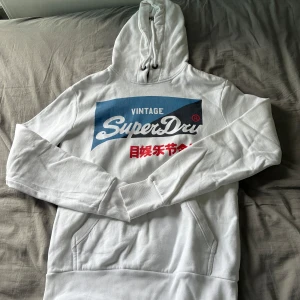 Vit Superdry hoodie storlek M - Vit hoodie från Superdry med stort blått och rött tryck på bröstet. Klassisk känguruficka, snörning i huvan och ribbade muddar. Skön och avslappnad passform, perfekt för chill dagar. Materialet känns mjukt och är troligtvis bomull.