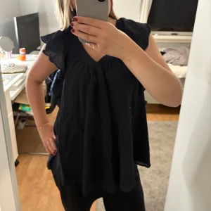 Mörkblå/svart blus från COS - Säljer en mörkblå nästan svart blus med v-ringning och volangdetaljer på axlarna. Blusen är ärmlös och har en luftig, loose passform som gör den superbekväm. Perfekt för dig som gillar en enkel men ändå söt stil.