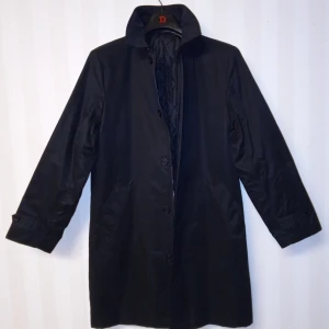 Svart rock med Dressmann märke, stl 48 - Klassisk svart rock från Dressmann med snygg krage och knäppning framtill. Insidan är quiltad för extra värme och komfort. Jackan har två fickor på sidorna och en innerficka. Perfekt för dig som vill ha en stilren och tidlös look.                                                     Kan skickas med extra pris via post.