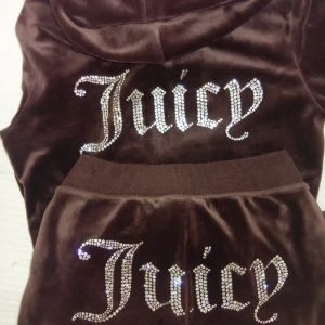 Juicy Couture velourset - Säljer ett Super trendigt velourset från Juicy Couture med zip hoodie och byxor. (Kan även tänka mig att byta mot ett grönt set) Båda delarna har stora glittriga Juicy-logos i strass på baksidan. Hoodien har dragkedja med J-berlock och snörning i luvan. Perfekt för dig som älskar 2000-talsvibbar och comfy stil. Setet är i bra skick och alla metalldelar och sånt är kvar, säljer det för ett bra pris och det går inte att pruta ner. Byxorna är i strl XXS och tröjan är i strl S.