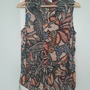 Mönstrad ärmlös blus - Säljer en ärmlös blus med coolt djur- och bladmotiv i svart, beige, orange och blått. Blusen har krage och knappar framtill, perfekt för dig som gillar unika prints och vill sticka ut. Tunn och luftig, passar grymt till jeans eller shorts.