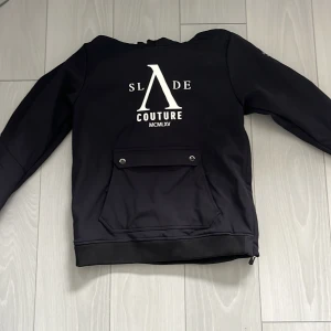 Svart skidjacka från Slade Couture - Svart skidjacka från Slade Couture med stor vit logga på ryggen och bröstet. Jackan har huva, stor ficka framtill med lock och tryckknappar samt mjukt foder. Snygg och stilren design som passar perfekt i skidbacke. Den har även en inbyggd munskydd/buff som täcker mun och näsa. Använd samanlangt 10 dagar skidåkning 