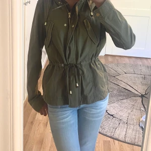 grön parkajacka med snörning - Snygg militär grön parkajacka med huva, dragkedja och knappar framtill. Jackan har snörning i midjan för en schysst passform och två fickor med lock på bröstet. Perfekt för  chill vår- eller höstdagar. St 38 men passar mig som har xs