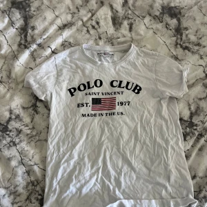 Vit t-shirt från Polo Club - Vit t-shirt från Polo Club Saint Vincent med tryck framtill i svart och amerikansk flagga. Klassisk rund hals och korta ärmar. Skön och lätt i bomull, perfekt till jeans eller shorts.
