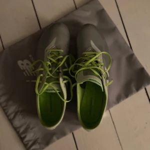 Silvriga New Balance fotbollsskor elite - Snygga fotbollsskor från New Balance i metallic silver med limegröna detaljer och snörning. Skorna har en cool kromad sula och gröna dobbar för grymt grepp på planen. Perfekt för dig som vill sticka ut med stil och prestanda.