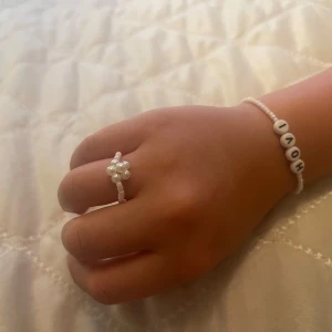 Pärlring och armband med textpärlor - Säljer ett sött set med en vit pärlring och ett matchande armband med små vita pärlor och bokstavspärlor som bildar ordet 'HOV1'. Ringen har en blomformad detalj av pärlor. Perfekt för dig som gillar unika och personliga smycken.