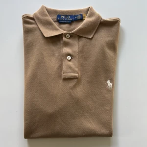 Beige pikétröja från Polo Ralph Lauren - Snygg beige pikétröja från Polo Ralph Lauren med klassisk krage och två knappar framtill. Tröjan har det ikoniska broderade logotypen i vitt på bröstet och är tillverkad i mjuk bomull. Perfekt för en clean och stilren look.