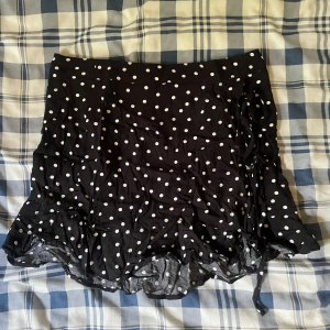 Svart prickig kjol från Pull&Bear - Supersöt svart kortkjol från Pull&Bear med vita prickar och volangkant. Kjolen har snörning på sidan som ger en extra snygg detalj. Perfekt för dig som gillar en lekfull och trendig stil.