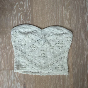 Vit stickad bandeau tubtopp - Supersöt vit stickad bandeau tubtopp med spetsig kant upptill och vackert hålmönster. Perfekt för sommardagar eller festivaler, lätt att matcha med höga jeans eller kjol. Mjuk och stretchig för skön passform.