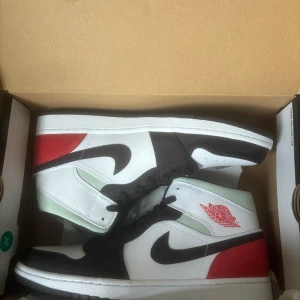 Nike Air Jordan 1 Mid svart/vit/röd - Nike Air Jordan 1 Mid sneakers med svart, vit och röd färgkombination. Klassisk svart swoosh, röd häl och Air Jordan-logga på sidan. Ovandel i skinn och mocka med svarta snören och perforerad tå. Snygg och ikonisk siluett som sticker ut.