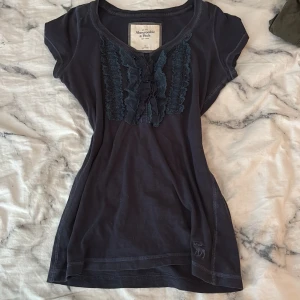 Mörkblå blus med spets från Abercrombie - Supersöt mörkblå blus från Abercrombie & Fitch med korta ärmar, v-ringning och dekorativa spetsvolanger framtill. Liten broderad älg nertill. Mjuk bomullskvalitet och figurnära passform. 