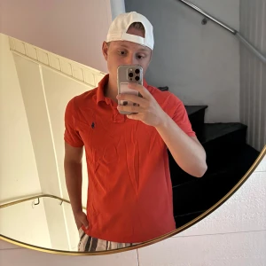 Röd pikétröja från Polo Ralph Lauren - Säljer en klassisk röd pikétröja från Polo Ralph Lauren med blå logga broderad på bröstet. Tröjan har krage, knappar vid halsen och är kortärmad. Perfekt för sommaren och enkel att matcha med shorts eller jeans. Sitter mer som M