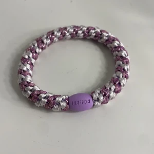 Lila och silver armband från KNEKKI - Säljer ett flätat armband från KNEKKI i lila och silver. Armbandet har ett coolt randigt mönster och en lila pärldetalj med märkesnamnet ingraverat. Perfekt accessoar för att ge din outfit lite extra färg och stil.