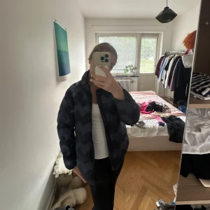 Svart och mörkblå pufferjacka med vågmönster - Säljer en riktigt snygg pufferjacka i svart och mörkblått med vågigt mönster. Jackan har hög krage, dragkedja framtill och en oversized passform som ger en cool vibe. Perfekt för kalla dagar när du vill sticka ut lite extra.