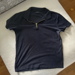 Svart pikétröja med dragkedja SCW - Svart pikétröja från SCW i storlek M. Tröjan har klassisk krage och en snygg guldfärgad half zip-dragkedja framtill. Kortärmad modell i mjukt bomullsmaterial, perfekt för en clean och stilren look.