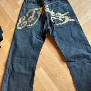 Ed Hardy jeans  - Mörkblå jeans från Ed Hardy med stort guldigt logotryck över bakfickorna. Dessa kan även spännas där bak! De är herr modell men jag som tjej har använd dessa och de sitter fint!. Dessa är väldigt unika då de har guld där bak!