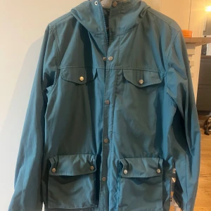 Blå vindjacka från Fjällräven M - Säljer en blå vindjacka från Fjällräven i storlek M. Jackan har huva, fyra stora fickor med tryckknappar och klassisk Fjällräven-logga på ärmen. Perfekt för uteliv och äventyr, tillverkad i slitstark polyester och bomull.