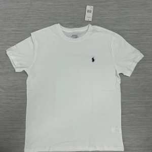 Vit t-shirt från Polo Ralph Lauren - Klassisk vit t-shirt från Polo Ralph Lauren med broderad marinblå logga på bröstet. T-shirten har rund halsringning och korta ärmar. Tillverkad i mjuk bomull för en clean och stilren look.