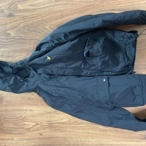 Svart vindjacka från Lyle & Scott - Svart regn och vindjacka från Lyle & Scott med huva, dragkedja och stora fickor framtill. Jackan har den klassiska gula loggan på bröstet. Perfekt för regn och blåsiga dagar och enkel att matcha med det mesta.