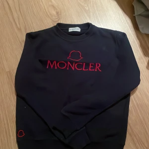 Svart Moncler sweatshirt med röd logga - Svart sweatshirt från Moncler med röd broderad logga och text på bröstet. Klassisk rund halsringning och långa ärmar. Tröjan har en clean look med en extra liten röd logga vid nederkanten. Perfekt för dig som gillar stilrena designer med en lyxig touch.