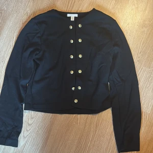 Svart kofta med guldknappar H&M - Svart kofta från H&M i storlek S med snygga guldknappar framtill. Modellen är croppad och har lång ärm, perfekt för att lyfta en enkel outfit. Tillverkad i mjukt bomullsmaterial som känns skönt mot huden.