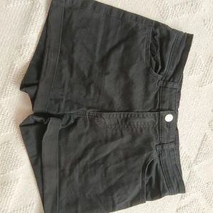 Svarta högmidjade jeansshorts - Svarta shorts i jeansmaterial med hög midja och klassisk femficksdesign. De har knapp och dragkedja framtill samt två bakfickor. Perfekta för en chill sommarlook eller när du vill ha något enkelt men snyggt.