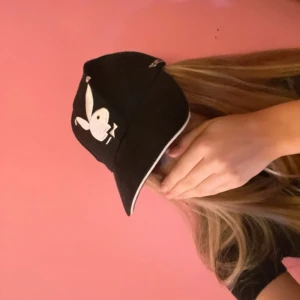 Svart keps från Playboy - Svart keps från Playboy med klassisk vit kaninlogga framtill och PLAYBOY-text broderad på sidan. Justerbar rem baktill för perfekt passform. Kepsen har böjd skärm och ventilerande hål upptill. Snygg och ikonisk accessoar för en cool streetstil.