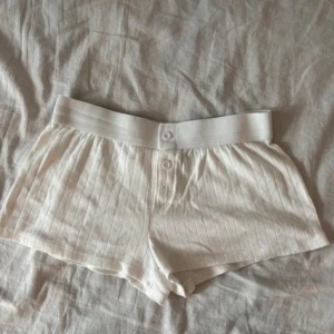 Brandy Melville Shorts - Säljer ett par vita shorts med ribbad struktur och bred resår i midjan. De har tre knappar framtill och är låga i midjan. Perfekta för chill dagar eller som mysplagg hemma. Materialet känns mjukt och stretchigt. Endast använda någon enstaka gång ❤️