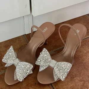 Beiga sandaler med strassrosett från Zara - Snygga beiga sandaler från Zara med smala remmar och låg klack. Skorna har en stor glittrig rosett framtill täckt av strass, vilket ger en extra glamorös touch. Perfekta för dig som vill sticka ut med detaljer och älskar bling.