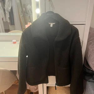 Mysig teddyjacka från H&M, mörkgrön - Teddyjacka från H&M i mörkgrönt, med dragkedja framtill och två stora fickor. Jackan har en hög krage och är tillverkad i mjukt fleece-material som ger en riktigt cozy vibe. Perfekt för kyliga dagar när du vill vara både varm och snygg.