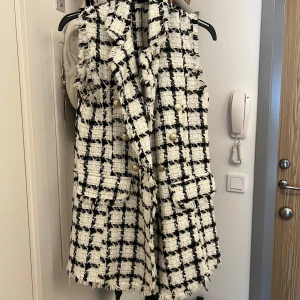 Rutig tweedväst med guldknappar - Snygg rutig väst i svart och vit tweed med råa kanter och dekorativa guldknappar. Västen har två fickor framtill och en klassisk krage. Perfekt för att lyfta vilken outfit som helst med en chic touch.