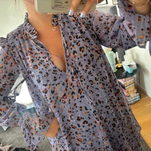 Lila leopardmönstrad kortklänning - Superfin lila kortklänning med leopardmönster i svart, orange och beige. Klänningen har volangdetaljer vid ärmslut och hals, knytning i halsen och långa, luftiga ärmar. 