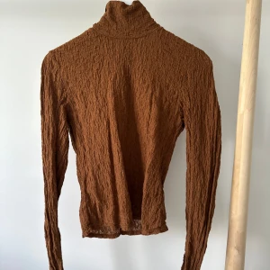 Brun polotröja med struktur - Säljer en brun polotröja med lång ärm och snyggt strukturerat tyg. Tröjan har en tight passform och hög krage, perfekt att styla med jeans eller kjol. Materialet känns lätt och stretchigt, vilket gör den bekväm att bära hela dagen.