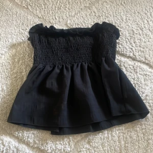 Svart volangig tubtopp - Supersöt svart tubtopp med smockad överdel och volangig nederdel. Toppen är axelbandslös och har en rynkad kant upptill. Perfekt att styla med jeans eller kjol.
