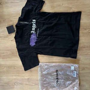 Svart t-shirt från Palm Angels - Säljer en svart t-shirt från Palm Angels med vit logga och lila tryck på bröstet. Klassisk passform och rund halsringning. Perfekt för dig som gillar streetwear och vill sticka ut med en ikonisk design.