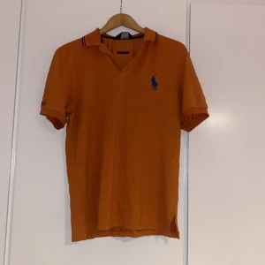 Orange pikétröja från Polo Ralph Lauren - Snygg orange pikétröja från Polo Ralph Lauren med blå logga på bröstet och diskreta ränder på kragen. Tröjan är i bomull och polyester, har korta ärmar och klassisk pikékrage. Perfekt för en sportig och avslappnad stil.