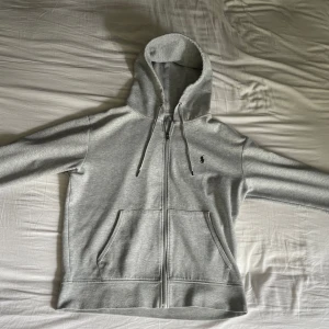 Grå hoodie från Polo Ralph Lauren - Snygg ljusgrå hoodie från Polo Ralph Lauren med dragkedja, huva med snörning och klassisk broderad logga på bröstet. Tröjan har känguruficka och är tillverkad i mjuk bomullsmix. Perfekt för chill dagar eller när du vill ha en clean look.