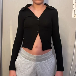 Svart ribbad croppad kofta - Svart croppad kofta med lång ärm och snygg krage. Tröjan är ribbad och har knappar framtill. Perfekt för dig som gillar en stilren och trendig look. Materialet är stretchigt och sitter tajt mot kroppen.