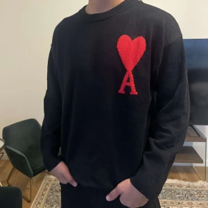 Ami sweater - Svart stickad tröja från Ami Paris med ikoniskt rött hjärta och A på bröstet. Klassisk rund halsringning, ribbade muddar och lång ärm. Perfekt för dig som vill ha en stilren men ändå unik look. Storlek S