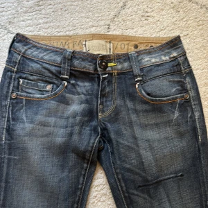 G-Star Raw jeans med broderade bakfickor - Snygga mörkblå jeans från G-Star Raw med coola broderade detaljer på bakfickorna och slitna effekter. Klassisk femficksmodell med orange kontrastsömmar och raka ben. Perfekta för dig som gillar streetstyle och vill ha ett par unika jeans. Nypris runt 1000kr och knappt använda. Hör av er vid frågor! 