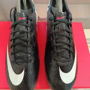 Nike Mercurial svarta fotbollsskor - Svarta Nike Mercurial fotbollsskor med vit swoosh och stora vita NIKE-bokstäver på hälen. Skorna har snörning, texturerad syntetisk ovandel och röda dobbar. Perfekta för dig som vill sticka ut på planen med stilren och sportig look.