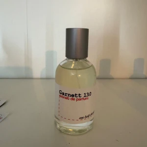 Garnett 13.0 Extrait de Parfum - En stilren parfymflaska med transparent glas och silverfärgad kork. Etiketten är vit med svart och röd text där det står Garnett 13.0 extrait de parfum. Doften känns modern och flaskan har en minimalistisk design som passar perfekt på hyllan.