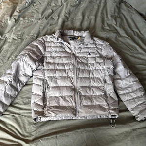 Ljusgrå dunjacka Polo Ralph Lauren M - Säljer en ljusgrå dunjacka från Polo Ralph Lauren. Jackan har quiltad design, dragkedja framtill, två sidofickor med dragkedja och justerbar dragsko nertill. Klassisk krage och diskret logga på bröstet. Perfekt för kyliga dagar.