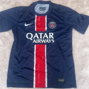 PSG hemmatröja 2024/25 - Mörkblå Paris Saint-Germain fotbollströja med röd och vit rand i mitten, Nike-logga och PSG-emblem på bröstet. Qatar Airways sponsortryck på framsidan. Klassisk passform och kortärmad modell i lätt och ventilerande material. Pris kan diskuteras vid snabb affär 